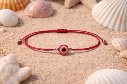 Red cord bracelet with a evil eye charm on sand with seashells