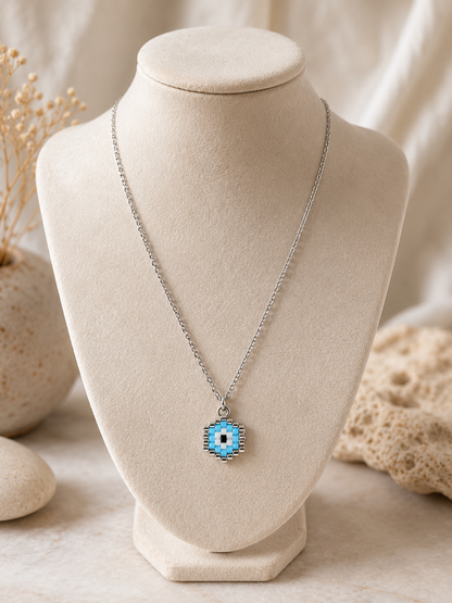 A silver chain necklace with a blue and white evil eye pendant, displayed on a beige fabric jewelry bust.
