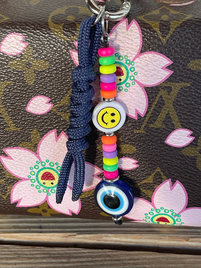 A multicolored bag charm featuring a smiley face and an evil eye design with colorful rubber beads on a navy blue tassel, displayed against a floral patterned background.
