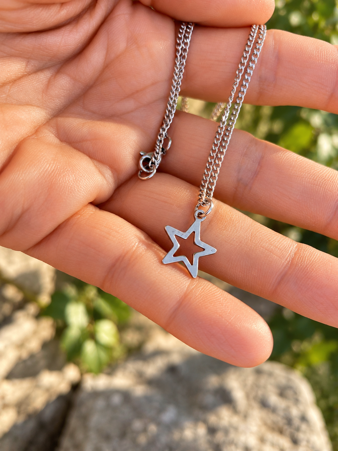 A silver-toned star-shaped pendant necklace with a curb chain, held in a person's hand against a blurred outdoor background of green foliage and rocks.
