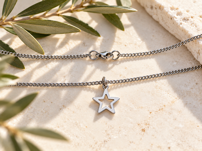A silver-toned two-layer necklace with a small open star pendant, displayed on a textured beige stone surface with olive branches.