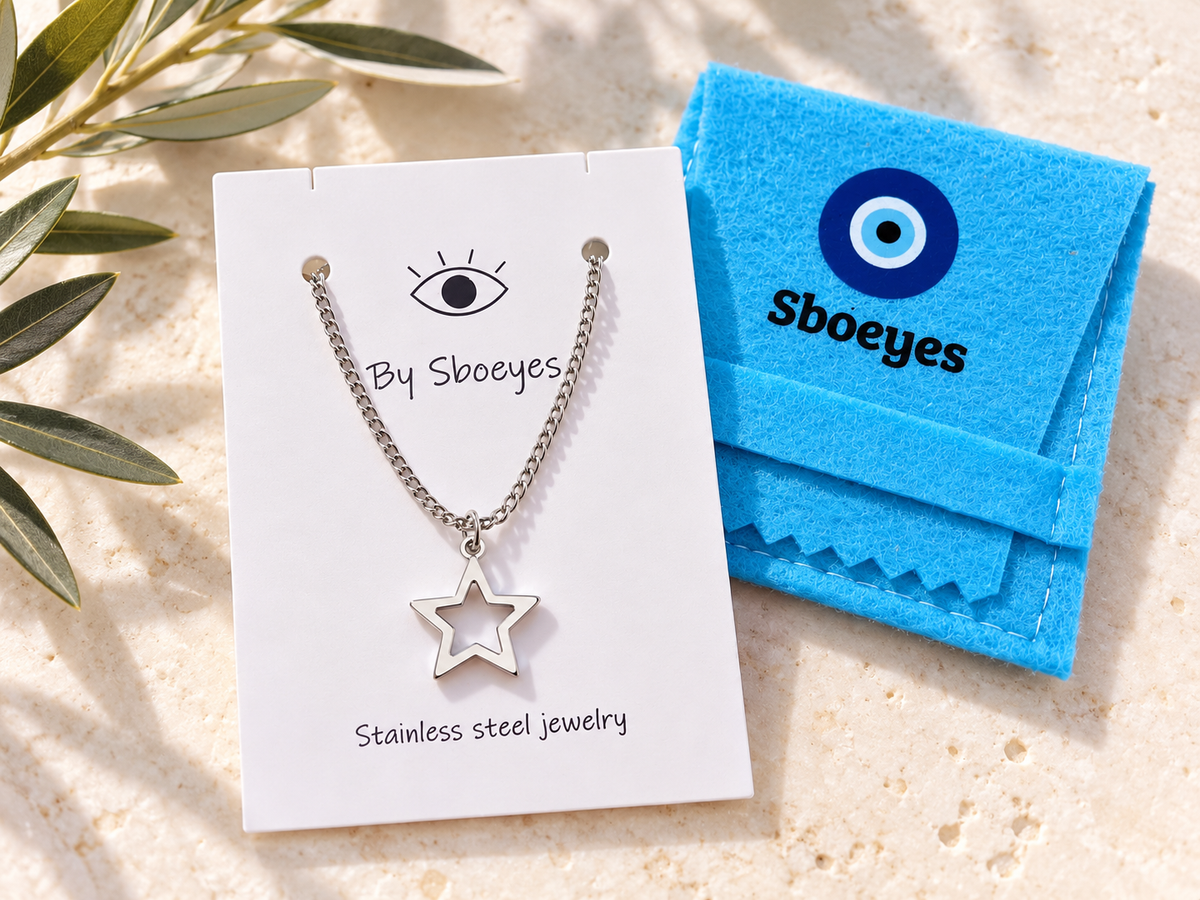 A silver star pendant necklace on a white display card next to a bright blue felt jewelry pouch branded Sboeyes, set on a light stone surface with olive leaves in the corner.