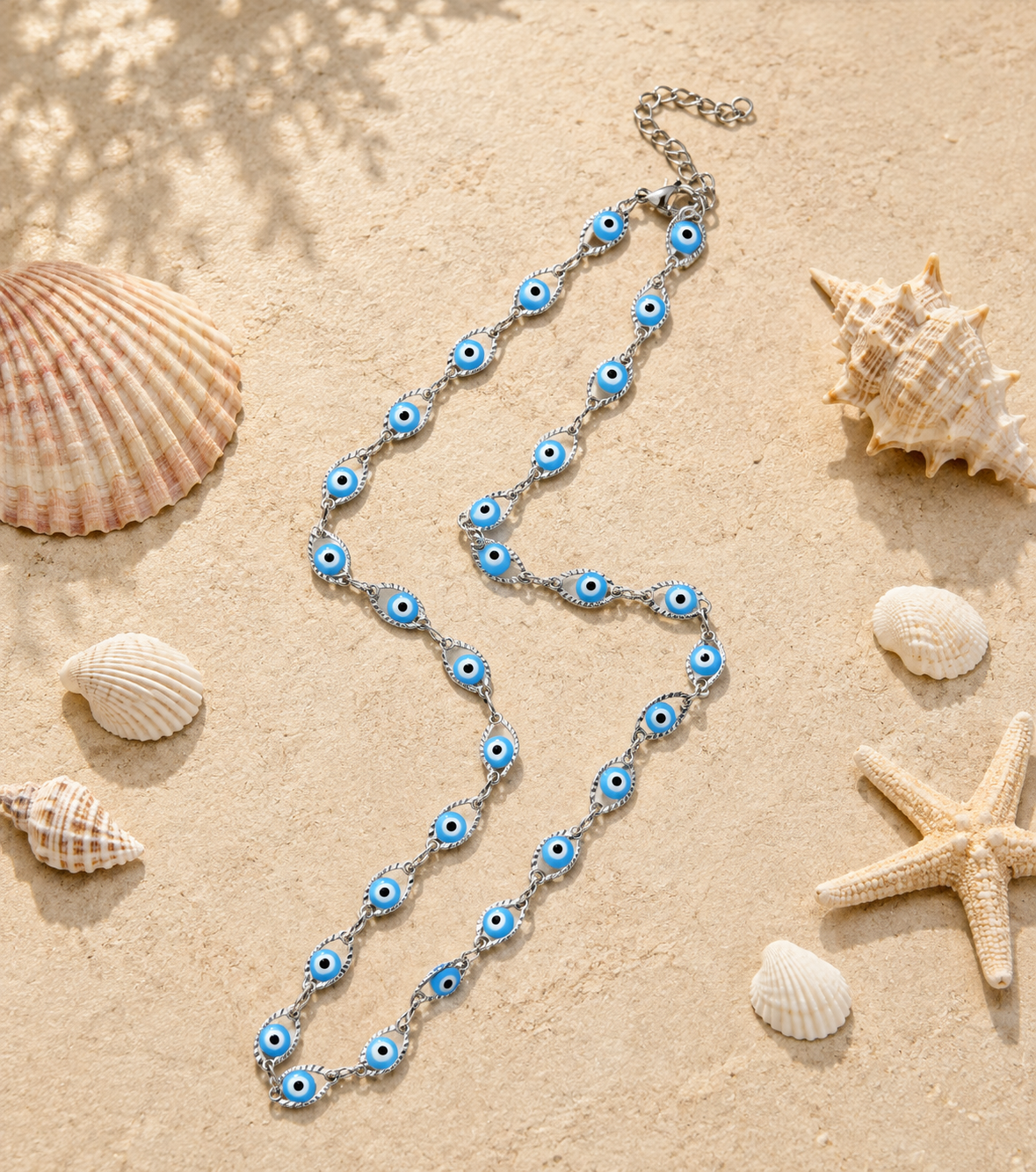A silver-toned chain necklace with blue and white evil eye charms, styled on a sandy beach background with seashells and a starfish.