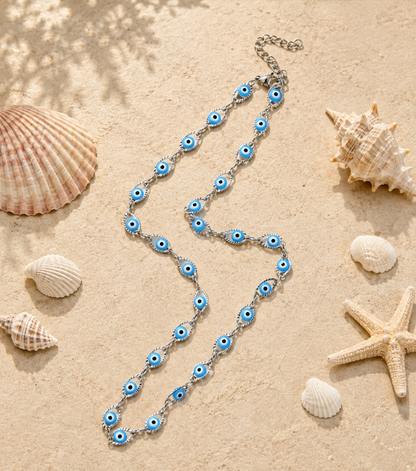 A silver-toned chain necklace with blue and white evil eye charms, styled on a sandy beach background with seashells and a starfish.