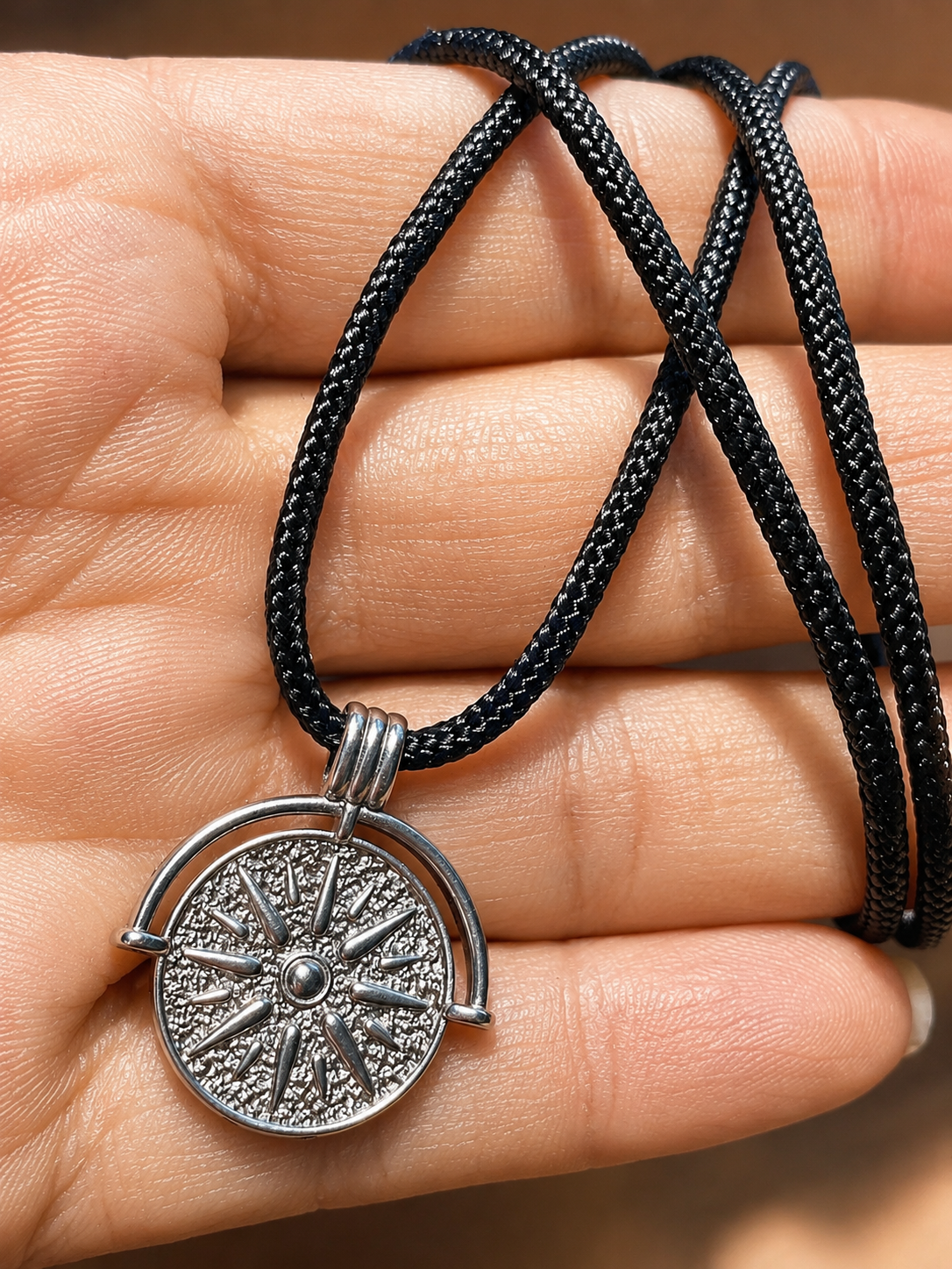 A close-up of a hand holding a black braided cord necklace with a silver-toned compass pendant.