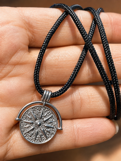 A close-up of a hand holding a black braided cord necklace with a silver-toned compass pendant.
