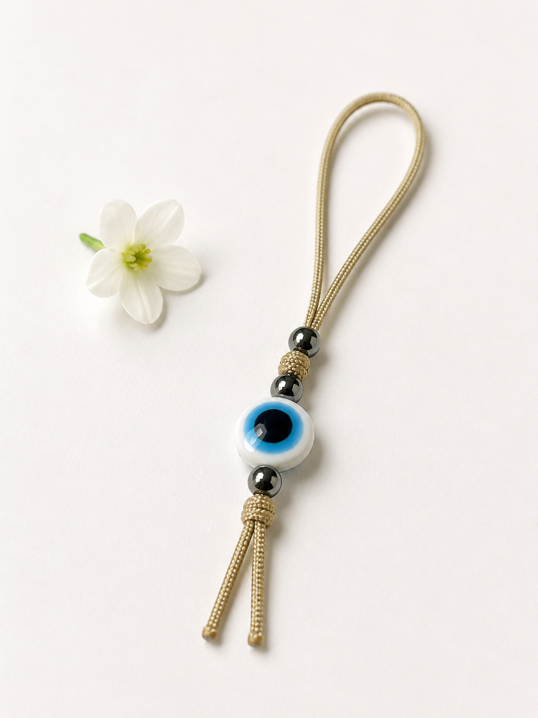 A gold-toned woven cord keychain or bag charm with a white evil eye bead, black spacer beads, and a tassel, photographed on a clean white background with a small white flower prop.