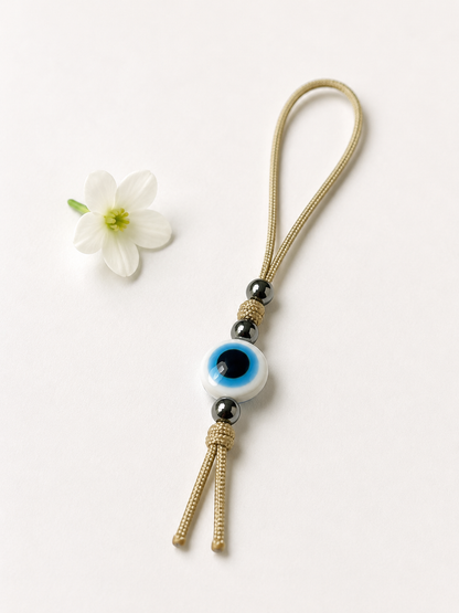 A gold-toned woven cord keychain or bag charm with a white evil eye bead, black spacer beads, and a tassel, photographed on a clean white background with a small white flower prop.