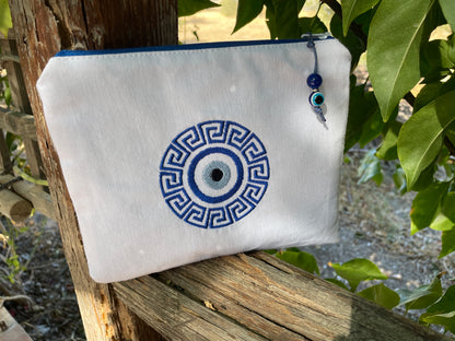 White pouch with blue decorative design hanging on a wooden post with greenery in the background