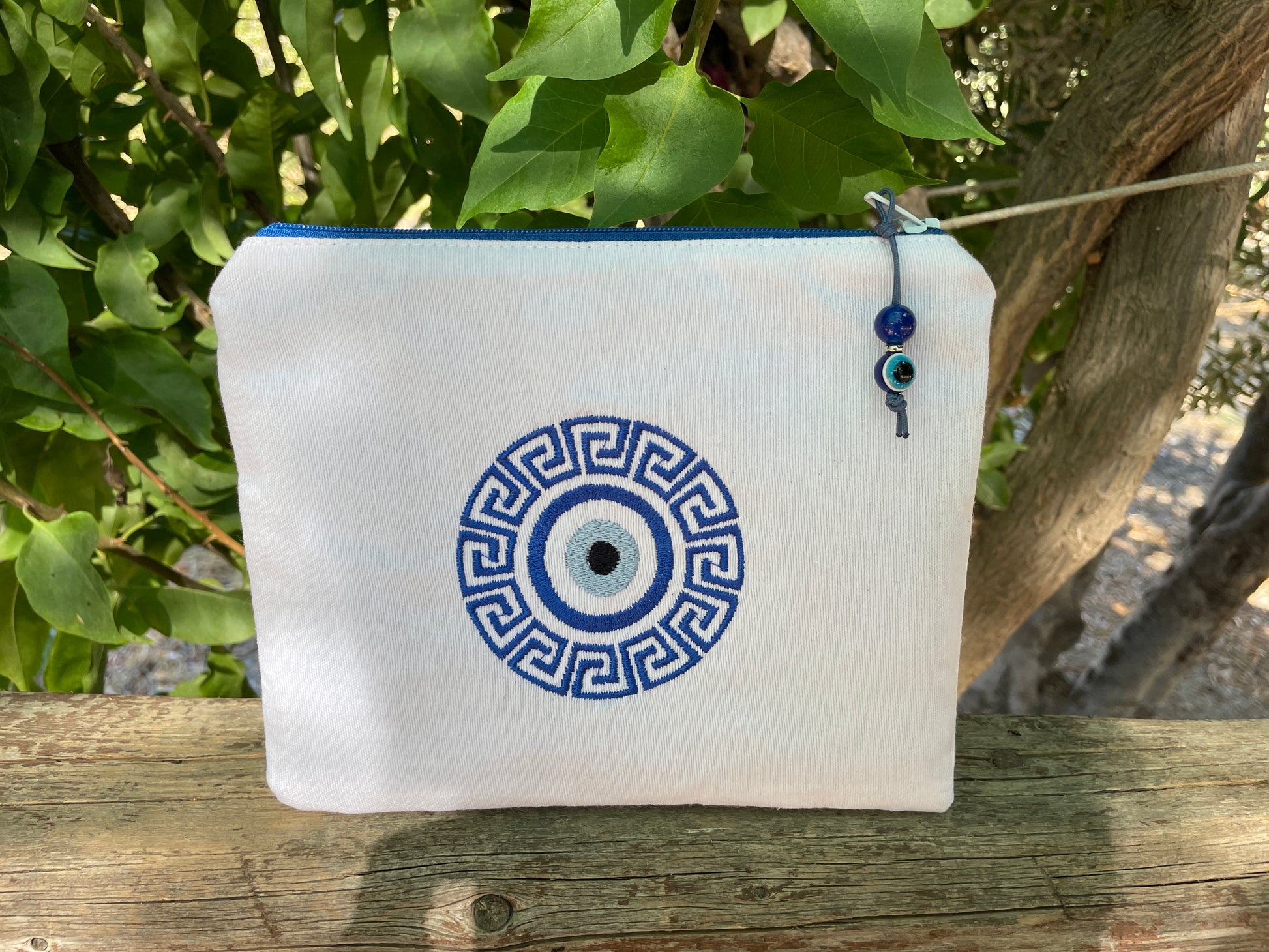 White pouch with blue and black decorative design on a wooden surface with greenery in the background