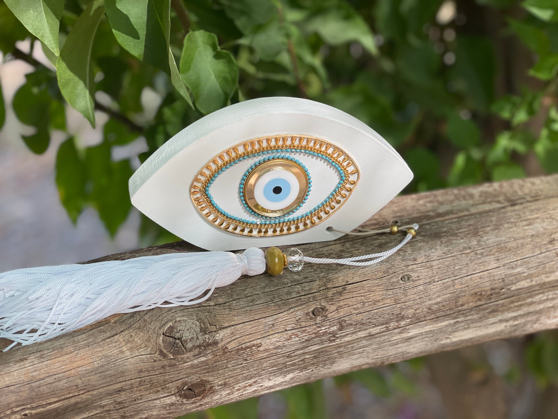 Decorative eye-shaped ornament with tassel on a wooden surface with green foliage in the background