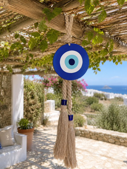 A blue and white evil eye hanging ornament with a beige rope tassel hangs from a wooden pergola under green vines, with a Mediterranean patio and sea in the background.