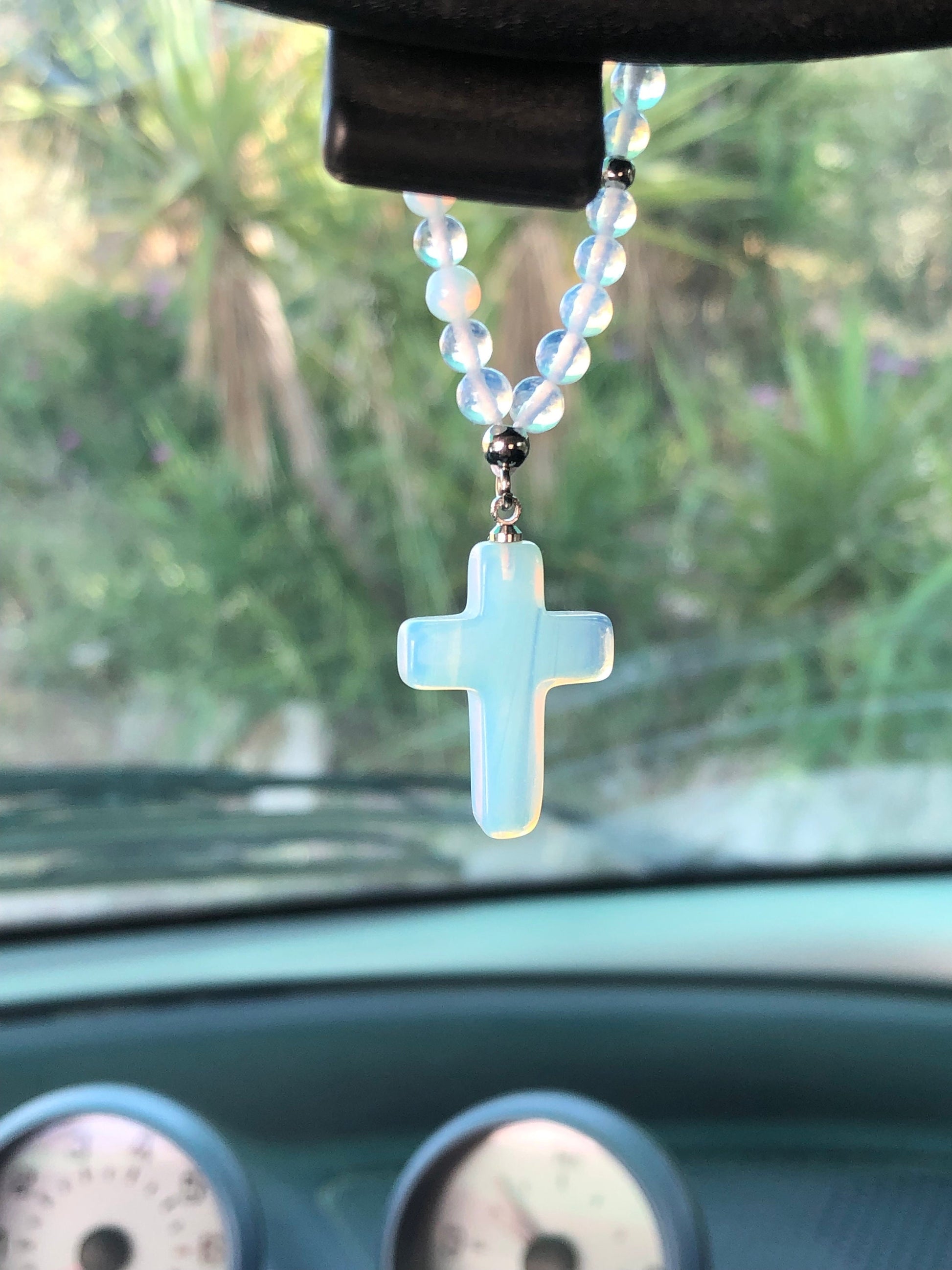 Moonstone cross car charm hangs from a beaded necklace, featuring soft blue beads and a translucent cross pendant.
